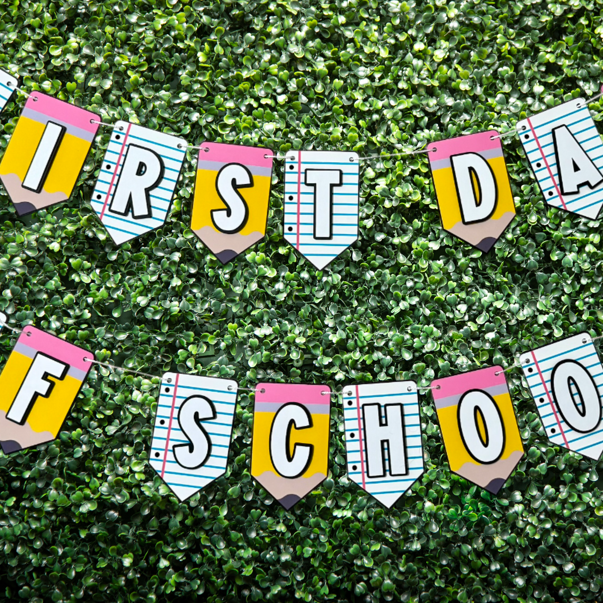 Colorful 'First Day of School' banner with pencil and notebook design on a green grass background.