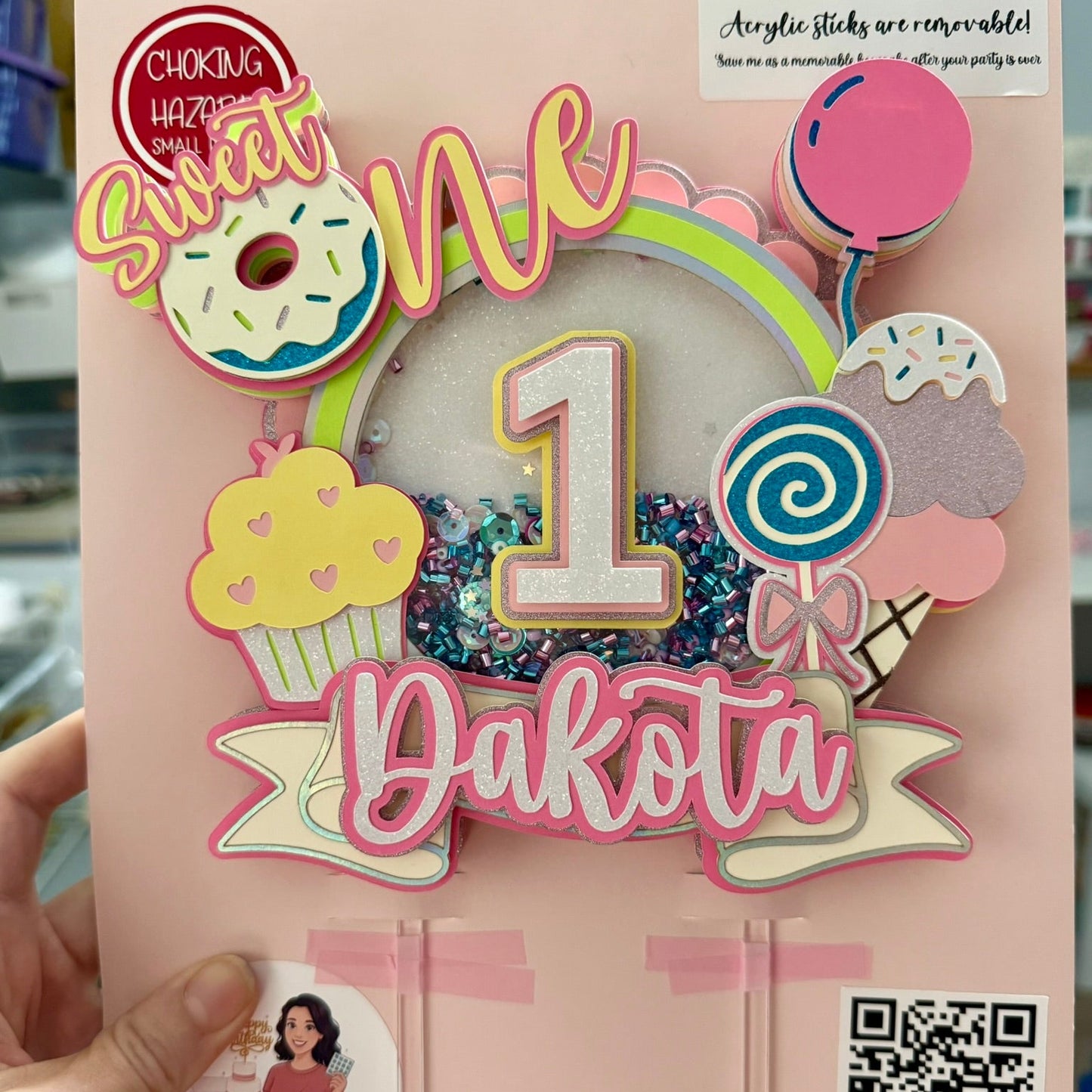 Decorative birthday cake topper with 'Sweet One' and 'Dakota' on a pink background for a first birthday celebration.