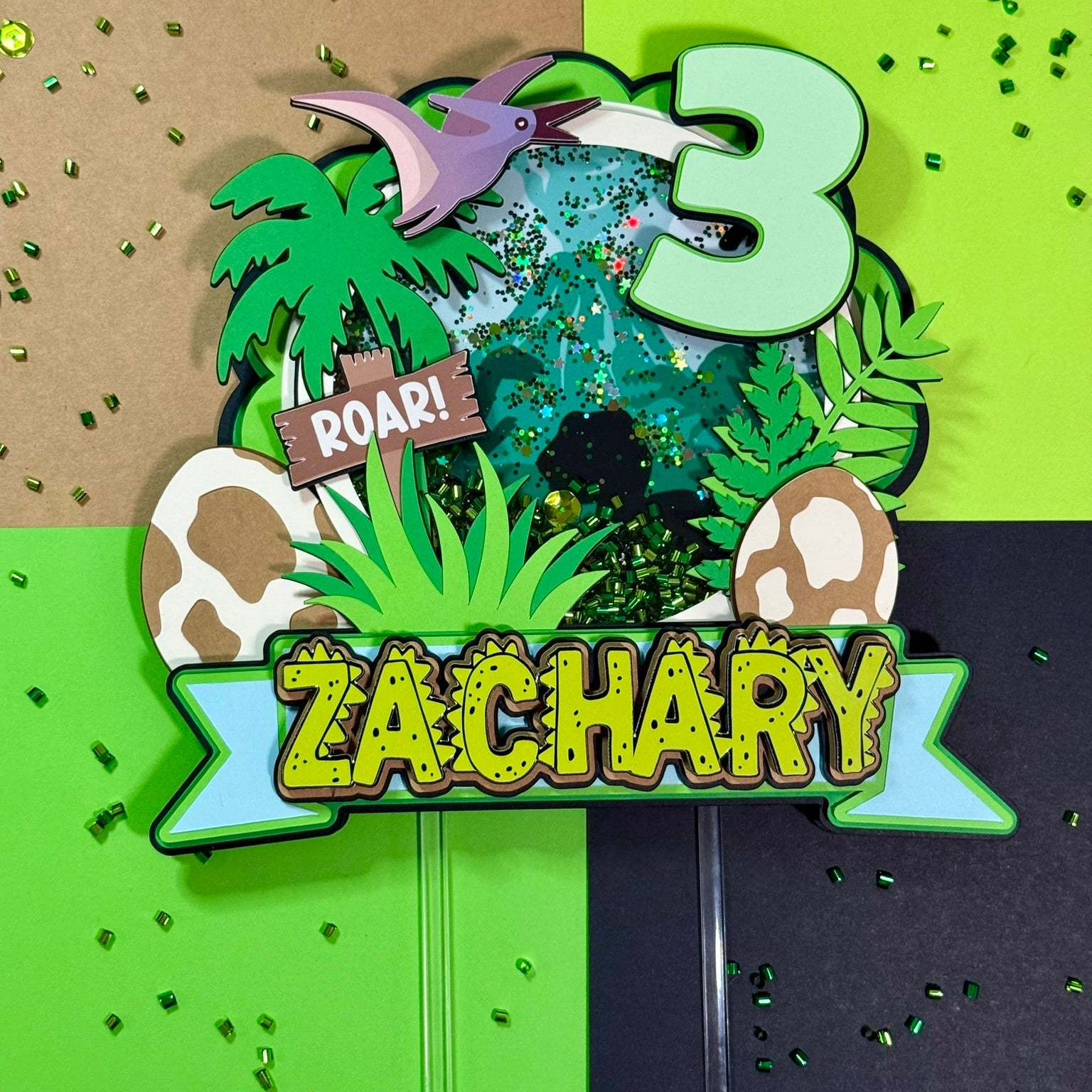 Dinosaur-themed birthday cake topper with number 3 and name 'Zachary' on a green and brown background.